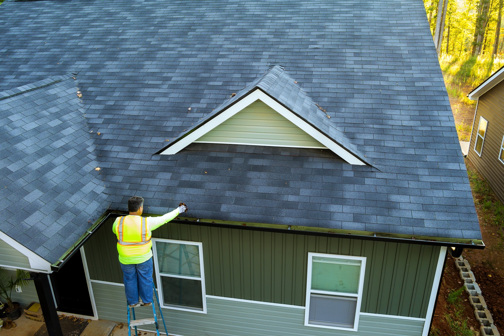 Using a ladder, worker cleans debris, dirt, fallen leaves from clogged roof gutter drain